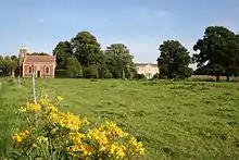 A grassy field, its uneaven surface barely discernable, is bright green under a blue summer's sky.  In the foreground are the bright yellow flowers of Rattle.  A small brick chapel stands in the rear left corner of the field, and beyond that are a row of mature oaks.  A gap in the row of trees gives a view of the frontage of Stainfield Hall