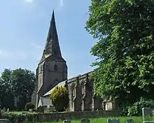 A stone church seen from the southeast, with a broach spire at the west end