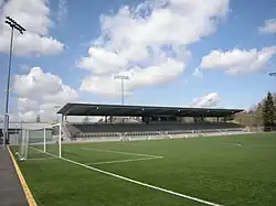 A grass field with some overhead lighting, a soccer goal, and stadium bleachers on one side.