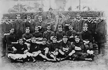 A black and white photograph of 27 men arranged in four rows posing for a team shot. The players in the front rows are in black jerseys and white shorts, while those not playing are in suits to the back.