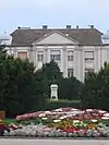 A colour photograph of Kálmán Tóth's memorial, front centre, before it several wreaths and flowers, surrounded by evergreen trees, and in the background a light stone building with Georgian escarpment