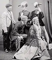 Five people in stage costumes; three are standing behind two who are seated