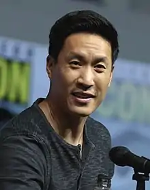 A handsome Asian man with short black hair smiles toward the camera while sitting behind a desk. A woman with short blonde hair sits next to him laughing. There are microphones on the desk in front of them.