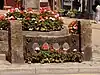 Stocks, made from central dark wooden stock beams with two holes in. The beams are supported by stone pillars. They have flowers planted in front of them, and the market cross, also with flowers, sits behind.