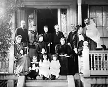 Late 19th century photo of a family of ten adults and four children posing on the front porch of a large house. The women wear long dresses with high collars and leg-o-mutton sleeves. The men have flowing moustaches. Marjory is a toddler in a long dress and black stockings.