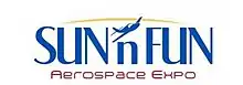 Logo depicts a small plane flying towards the viewer and facing the left. The background consists of two concentric circles (inner is yellow depicting the sun, outer is light blue to represent the sky). On the right is a palm tree and on the left (above the plane) is the name "SUN 'n FUN".