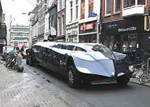 A dark-blue stretch-limousine-type vehicle in a narrow shopping street