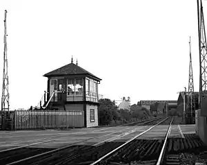 Raised cabin on left of railway lines stretching into distance on a left curve with large commercial buildings behind