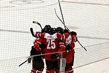 A group of hockey players stand on the ice in a circle, hugging each other and celebrating. They are all wearing red and white sweaters.