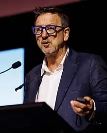 Paul Pritchard in blue suit and blue spectacles speaking at a podium.