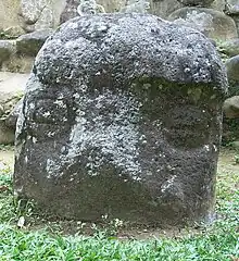 A low standing stone protruding from short grass, with the stonework of a structure visible behind it. The upper portion of the stone has a raised area perhaps representing a cap or helmet. Below this are carved two eyes in the form of circles divided by a horizontal line, giving the appearance of being closed. The cap and the area between the eyes are covered with pale lichen