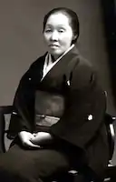 A Japanese woman of middle age, wearing a dark kimono, seated, with hands folded in her lap.