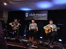 Talisk three men sitting on an indoor stage performing with the fiddle, concertina, and guitar