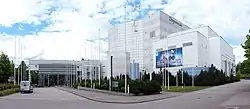 External view of building clad in white and silver siding, surrounded by flagpoles and greenery