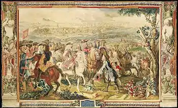 Tapestry shows a crowd of men mounted on horseback. They are dressed in early 18th century style with three-cornered hats and curly shoulder-length wigs. The man in the center rides a white horse, holds a sword and wears a metal cuirass. In front of him, a man on foot has his hat off and slightly bows. A walled fortress city is in the background.