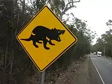 A square-shaped metal tilted at 45 degrees on a metal post. The sign is painted yellow with a picture of a black devil in profile. It is at the side of a straight road cutting through wooded forest and two vehicles can be seen.