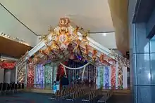 layers of colurful Māori carvings foming the meeting house with a raised wooden floor forming a small stage, carpet and chairs in the foreground. An adult and child are walking through the space.