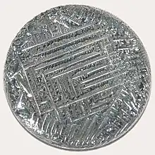 A shiny silver-white medallion with a striated surface, irregular around the outside, with a square spiral-like pattern in the middle