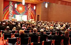 A ground-level view of the backs of around a dozen rows of chairs, filled with men and women, all facing the front of the room. A brightly colored artwork with a rainbow and sun design fills the facing wall. The center of the artwork holds an open Torah ark, and above it is a representation of the two tablets holding the Ten Commandments.