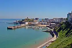 In the centre of the image is a wide, curved harbour with a sandy beach and a grassy cliff to the right. There are small boats moored in the harbour, and above the dark harbour walls are many pastel-coloured houses. Rising behind them to the left is a small hill with a tower.