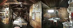 Dimly lit interior of a timber structure with a long table on which are papers and other unidentifiable objects. There are also bunks, sheles and packing cases. To the right, under a small window, is a smaller table with a chair.