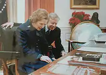  Margaret Thatcher seated behind the Resolute desk with Jimmy Carter looking over her shoulder as she read the plaque installed on the desk.