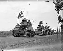 Photograph of several tanks and a truck driving along a road