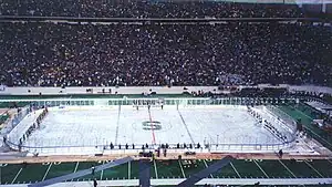 Two teams of many hockey players stand at either end of a rink built on top of a football field as thousands of people look on from the stands.