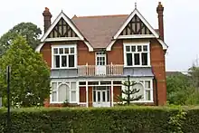 A detached red bricked building, with two chimneys, behind a green hedge and a tree to the left.