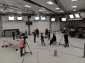 A bright white stage with actors in black suits and camera operators.