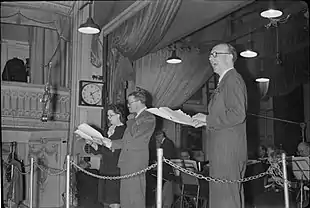 Photo taken at a recording session, with two performers, front, and band and conductor behind