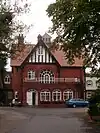 A red brick house, at the end of a long drive, seen from the south. Prominent is a gable with black and white striped pebbledash.