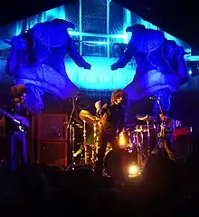 Three men standing on a stage in front of a crowd; two are holding guitars while the one of the center is holding a microphone. Audio equipment, a drum set, lighting, and other stage props can also be seen in the background.