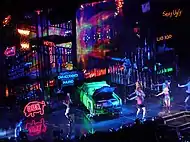 Bird's eye view of a stage, showing large scaffoldings, neon signs and a green car lying in the middle.