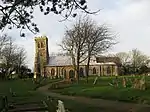 A stone church seen from the south, with a slightly leaning west tower on the left, a long south aisle to the right, and a porch