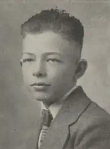 A white teenaged boy with dark hair; he is wearing a suit coat and tie, and is facing the camera's left.