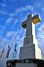 View of a large stone cross carved with the Croatian coat of arms. Outlined against the blue sky are the cross and three vertically hung flags.