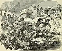 Several groups of black people are fighting in the tall grass with weapons. In the background of the image is a village with festive activities and what looks like a man practicing a ritual.