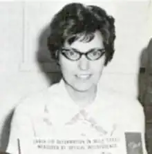 A white woman with short dark hair, wearing cat-eye glasses and a white blouse with a wide pointed collar