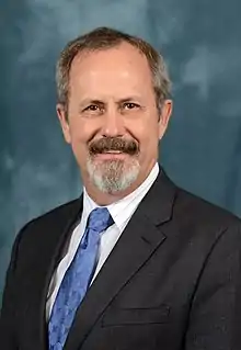 Photo of Caucasian male, wearing a grey suit with white shirt and red tie.