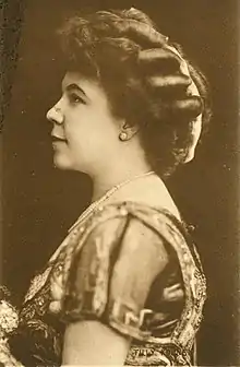 A white woman in profile, facing viewer's left; her dark hair is in an updo, and she is wearing a beaded and embroidered gown, a string of pearls, and an earring.