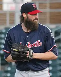 A man in a navy blue baseball jersey, gray pants, and a navy cap with hands held together in his black glove