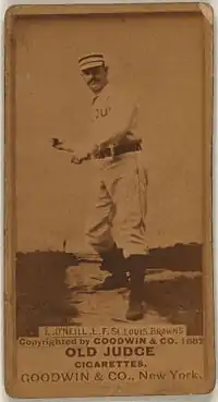 A sepia-toned image of a man wearing an old-style white baseball uniform and cap throwing a baseball