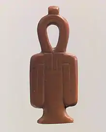 A red stone amulet shaped like a column with a looped top and two loops hanging at the sides