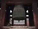 A large bronze bell with cross design hanging in an open roofed belfry.