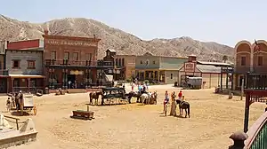 Squarish buildings made of wood and bricks surround a square, where horses and carriages gather.