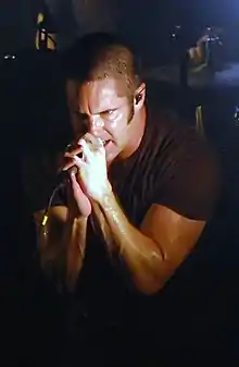 A man wearing a black shirt singing into a microphone, in front of a drum kit.