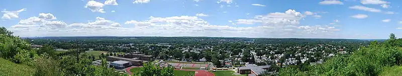 A wide panoramic shot begins and ends in bushes but is graced by two and three story city houses throughout most of the image. Front and center are three large schools featuring a track, tennis courts, and a football field. Behind the houses is a river flowing right to left.