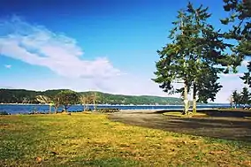 Picnic tables, empty parking lot, grass fields, and a lone tree with the water in the background