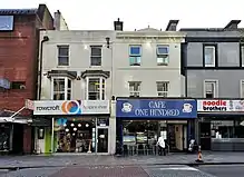 Four terraced buildings, three of them small, all two storeys, in a high street setting. The two central buildings are white; the building on the furthest left is of red brick, and the building furthest right is grey.